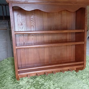 Puede incluir: Un estante de pared de madera de color marr&oacute;n medio con una cornisa superior plana, dos estantes horizontales y un riel inferior con seis peque&ntilde;os ganchos de madera orientados hacia abajo. El panel trasero tiene tablones verticales.