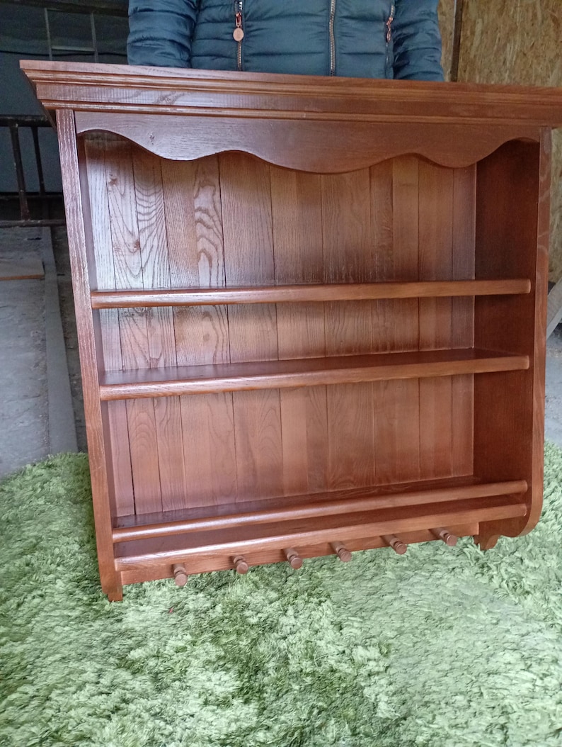 Puede incluir: Un estante de pared de madera de color marr&oacute;n medio con una cornisa superior plana, dos estantes horizontales y un riel inferior con seis peque&ntilde;os ganchos de madera orientados hacia abajo. El panel trasero tiene tablones verticales.