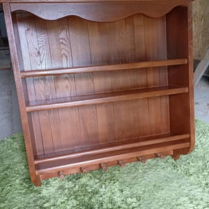 Puede incluir: Un estante de pared de madera de color marr&oacute;n medio con un borde superior festoneado, dos estantes horizontales y un riel inferior equipado con seis peque&ntilde;os ganchos de madera para colgar objetos. La unidad tiene un panel trasero de listones.
