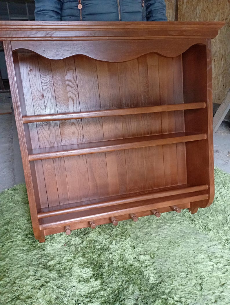 Puede incluir: Un estante de pared de madera de color marr&oacute;n medio con un borde superior festoneado, dos estantes horizontales y un riel inferior equipado con seis peque&ntilde;os ganchos de madera para colgar objetos. La unidad tiene un panel trasero de listones.