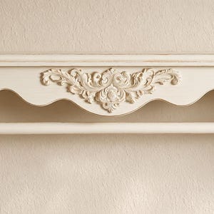 Könnte beinhalten: Ein cremefarbenes Wandregal mit gewellter Kante und einer zentralen, floralen Schnitzerei. Das Regal verfügt über eine horizontale Stange, die zwischen zwei seitlichen Halterungen befestigt ist und zum Aufhängen von Gegenständen wie Handtüchern dient.