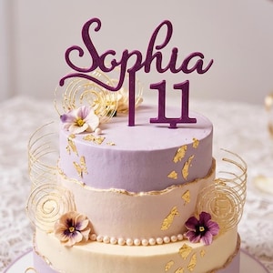 Könnte beinhalten: Dreistöckige Torte mit Lavendel- und Cremefrosting, verziert mit Blattgold und essbaren Blumen. Ein lila Glitzer-Tortentopper mit der Aufschrift "Sophia 11". Die Torte ist mit goldenen Spiralen verziert.
