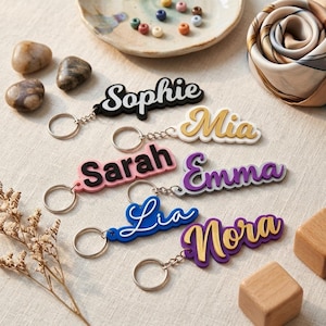 Op de afbeelding: Zes gepersonaliseerde naamsleutelhangers met gelaagde acryldesigns. De sleutelhangers tonen de namen Sophie in zwart, Mia in goud en wit, Sarah in zwart en roze, Emma in paars en wit, Lia in wit en blauw, en Nora in goud en paars.