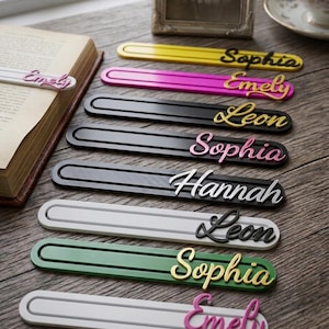 Peut inclure: Une collection de marque-pages rectangulaires, plats et allongés, de différentes couleurs dont jaune, rose, noir, blanc et vert. Chaque marque-page présente un nom en relief écrit en cursive dans une couleur contrastante, comme Sophia, Emely, Leon et Hannah.