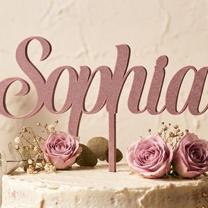 Op de afbeelding: Een oudroze glitter taarttopper met de naam Sophia in een cursief lettertype. De topper staat op een wit geglazuurde taart die is versierd met lichtpaarse rozen en kleine takjes groen.