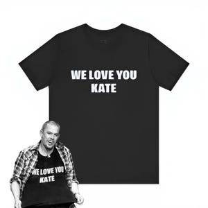 Può includere: T-shirt nero con la scritta bianca "WE LOVE YOU KATE". La t-shirt è mostrata su sfondo bianco. L'immagine mostra anche una persona che indossa la stessa t-shirt.