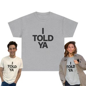 Könnte beinhalten: Ein hellgraues T-Shirt mit dem Text "I TOLD YA" in fetten schwarzen Buchstaben. Das Bild zeigt auch zwei Personen, die das gleiche T-Shirt tragen, eines in Weiß und das andere in Grau.