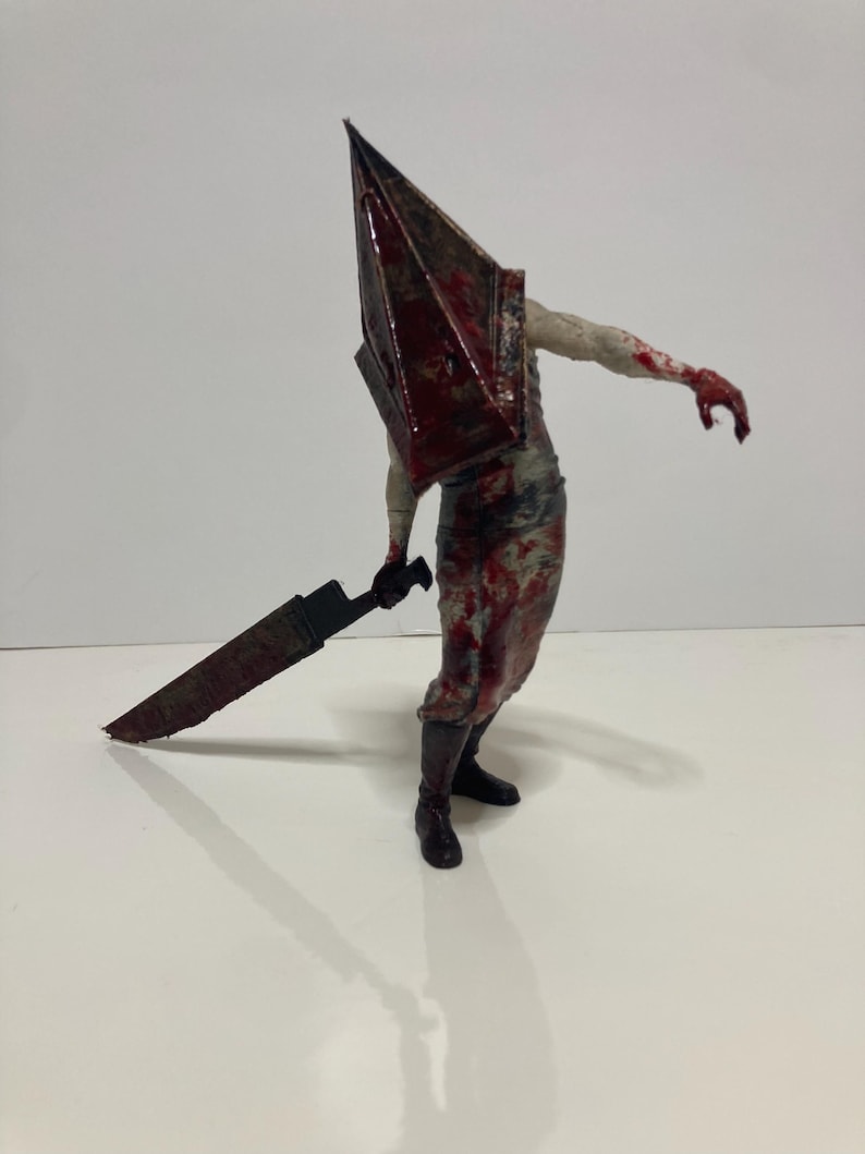 May include: A detailed figurine of a character with a large, blood-stained pyramid-shaped helmet. The figure is holding a large, rusty blade. The character is wearing a blood-splattered outfit and black boots. The overall aesthetic suggests a horror theme.