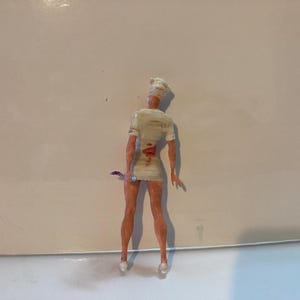 May include: A small figurine of a nurse, viewed from the back, wearing a white uniform with blood-like markings. The nurse is holding a small, curved blade. The figurine has a white cap and pale skin.
