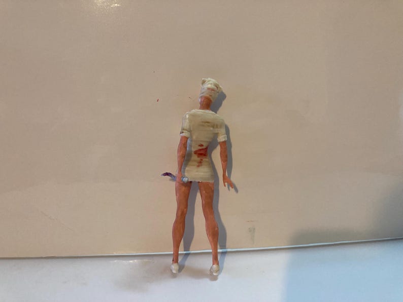 May include: A small figurine of a nurse, viewed from the back, wearing a white uniform with blood-like markings. The nurse is holding a small, curved blade. The figurine has a white cap and pale skin.