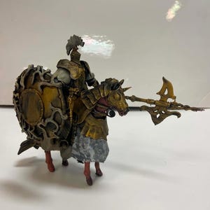 Op de afbeelding: Een miniatuurfiguur van een bereden ridder in verweerd goudkleurig en grijs harnas, rijdend op een paard met bijpassende gouden paardenharnas. De ridder draagt een groot, versierd rond schild en een lang, puntig wapen met gedetailleerde afwerking.