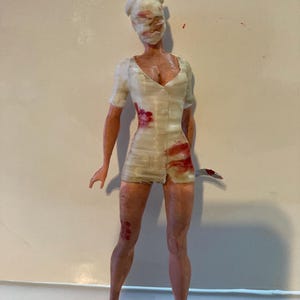 May include: A figurine of a nurse in a white uniform with blood-like stains and a knife. The nurse's face is obscured by a white mask. The figurine is standing on a white surface.