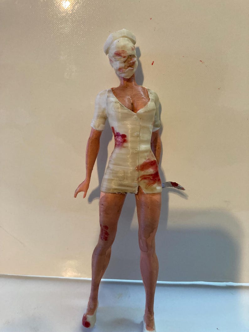 May include: A figurine of a nurse in a white uniform with blood-like stains and a knife. The nurse's face is obscured by a white mask. The figurine is standing on a white surface.