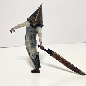 May include: A detailed figurine of a character from the Silent Hill video game series. The figure features a pyramid-shaped head, a white and gray outfit, and a large, rusty blade. The figure is posed with the blade held at its side.