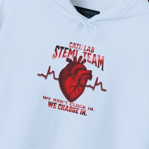Puede incluir: Sudadera con capucha blanca con bolsillo delantero y capucha con cordón. La parte delantera presenta el texto "CATI LAB STEM TEAM" encima de un gráfico de corazón rojo con una línea de latido. El texto "WE DON'T CLOCK IN. WE CHARGE IN." está debajo. La manga derecha tiene el texto "Lisa Smith DN."