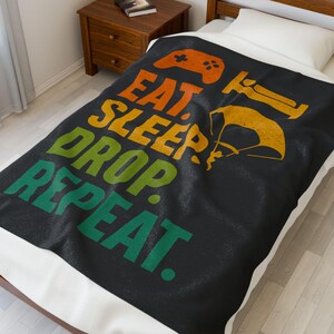 May include: Black blanket with the words "EAT SLEEP DROP REPEAT" in orange, yellow, green, and white. The blanket also features a video game controller and a parachute design in orange and yellow.