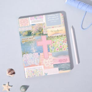 Può includere: Un diario decorativo con un design a collage con una croce rosa, motivi floreali e citazioni ispiratrici. Il diario include il testo di Isaia 66:13 e Proverbi 3:5-6. Sono visibili anche uno stilo bianco e filo blu.