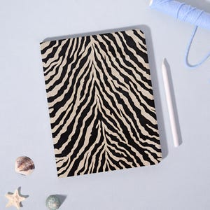 Pode incluir: Uma capa retangular para tablet com um padrão de riscas de zebra em preto e bege. A capa está posicionada ao lado de uma caneta stylus branca, um carretel de linha azul claro, uma pequena concha e uma pequena estrela-do-mar num fundo azul.
