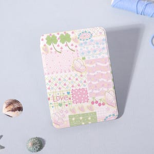 May include: A rectangular, pastel-coloured case with a patchwork design featuring pink, green, and white squares. The design includes butterflies, four-leaf clovers, cherries, and the word "Love".