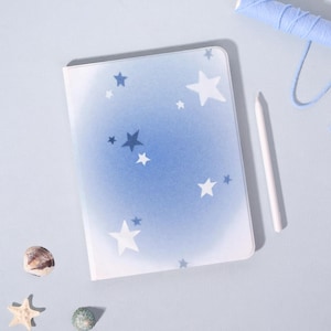 May include: A notebook with a blue gradient cover featuring white star designs. A white stylus, blue thread, a seashell, a starfish, and a small green stone are next to the notebook.