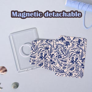 Könnte beinhalten: Eine Handyhülle mit einem floralen Muster in Marineblau und Creme, zusammen mit einer transparenten, rechteckigen Hülle und einem weißen Ringgriff. Der Text "Magnetic detachable" wird oben im Bild angezeigt.