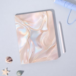 Op de afbeelding: Een parelmoeren tablet hoesje met een vloeiend, iriserend ontwerp in beige, goud en wit. Een witte stylus en een spoel blauw garen liggen naast de hoes. Schelpen en een zeester zijn ook zichtbaar.