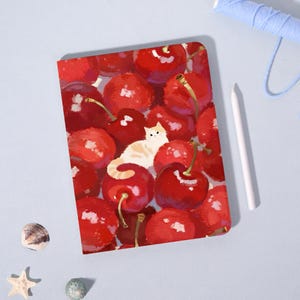 May include: A notebook featuring a vibrant illustration of ripe red cherries with green stems. A cream-colored cat with orange markings is nestled among the cherries. A white pen and blue thread are in the background.