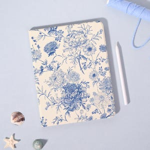 Può includere: Custodia per tablet color crema con motivo floreale blu con peonie e altri fiori. Uno stilo bianco e una bobina di filo blu sono accanto alla custodia. Il design è vintage e botanico.