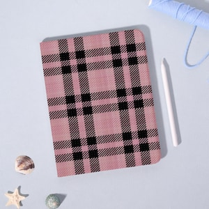 May include: A pink and black plaid notebook. The notebook has a rectangular shape with rounded corners. A white stylus and blue thread are next to the notebook. A seashell, starfish, and a small green stone are also in the image.