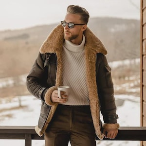 Puede incluir: Chaqueta de cuero marrón con cuello y forro de borrego beige. La chaqueta tiene cintura y puños con hebilla, y se lleva sobre un jersey de cuello alto blanco y pantalones marrones. La persona sostiene una taza blanca.