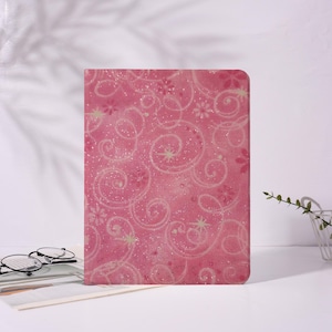 Könnte beinhalten: Ein rosa Notizbuch mit einem glitzernden Muster aus weißen Wirbeln, Sternen und Blumen. Das Notizbuch steht auf einer weißen Oberfläche, mit einer Brille und einer Zeitschrift im Vordergrund.