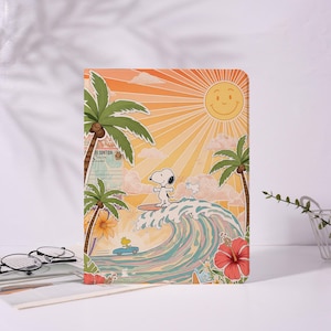 May include: A rectangular notebook featuring Snoopy surfing a wave, with a smiling sun and palm trees against an orange sky. A blue car with Woodstock is also depicted. The design has a vintage, cartoon style.