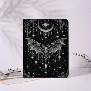 May include: Black notebook with a celestial bat design. The cover features a detailed illustration of a bat with outstretched wings, a crescent moon, stars, and decorative hanging chains. The design is in shades of gray and white against a black background.