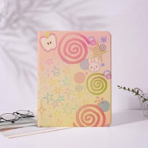 May include: A rectangular tablet case featuring a pastel gradient background decorated with various colorful illustrations including pink and green spirals, a small white rabbit face, a light-colored apple, and numerous multi-colored stars.