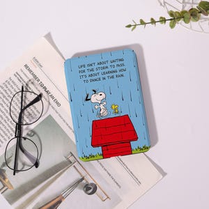 May include: A rectangular light blue case featuring a graphic of Snoopy and Woodstock dancing on a red doghouse in the rain. The text reads: LIFE ISN'T ABOUT WAITING FOR THE STORM TO PASS. IT'S ABOUT LEARNING HOW TO DANCE IN THE RAIN.