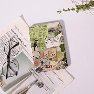 May include: A rectangular e-reader case featuring a collage of green and beige paper scraps, cartoon character illustrations, and Japanese text. The design includes small white bunny figures, a bear icon, and various graphic elements layered together.