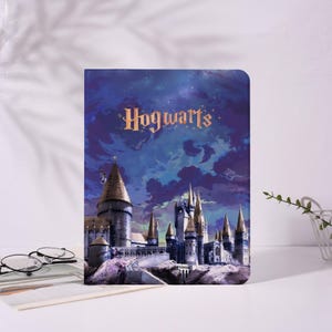 May include: A rectangular notebook featuring a detailed illustration of Hogwarts Castle under a starry night sky. The word "Hogwarts" is written in gold script across the top. The notebook is set against a white background.