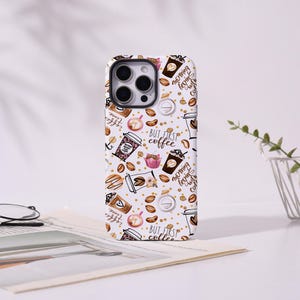 Op de afbeelding: Een witte telefoonhoes met een herhaald patroon van koffiekopjes, koffiebonen en de tekst "But first coffee". De hoes heeft koffie-thema illustraties in bruin, roze en goud.