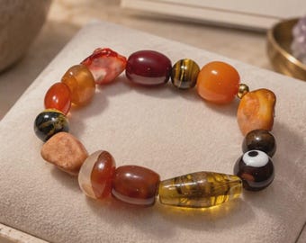 Könnte beinhalten: Ein Perlenarmband aus verschiedenen polierten Edelsteinen in Orange-, Rot-, Braun- und Gelbtönen, einschließlich einer dunklen Perle mit weißem augenähnlichem Motiv, kreisförmig angeordnet auf einem hellen Präsentationskissen.