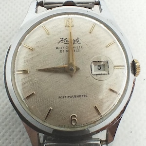 May include: A vintage silver wristwatch with a textured, light-coloured face. The watch has gold hands and markers, a date window showing the number 5, and the text "Kiehle Automatic 21 R 315 Antimagnetic". The watch strap is silver.