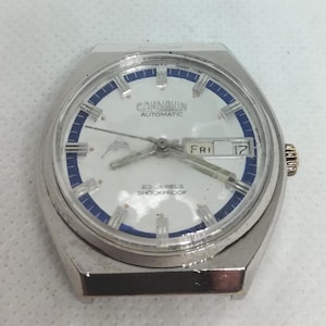 May include: A silver-toned Carnavin automatic wristwatch with a white face, blue accents, and a day-date display. The watch face features the text "CARNAVIN AUTOMATIC" and "22 JEWELS SHOCKPROOF". The day display shows "FRI 17".