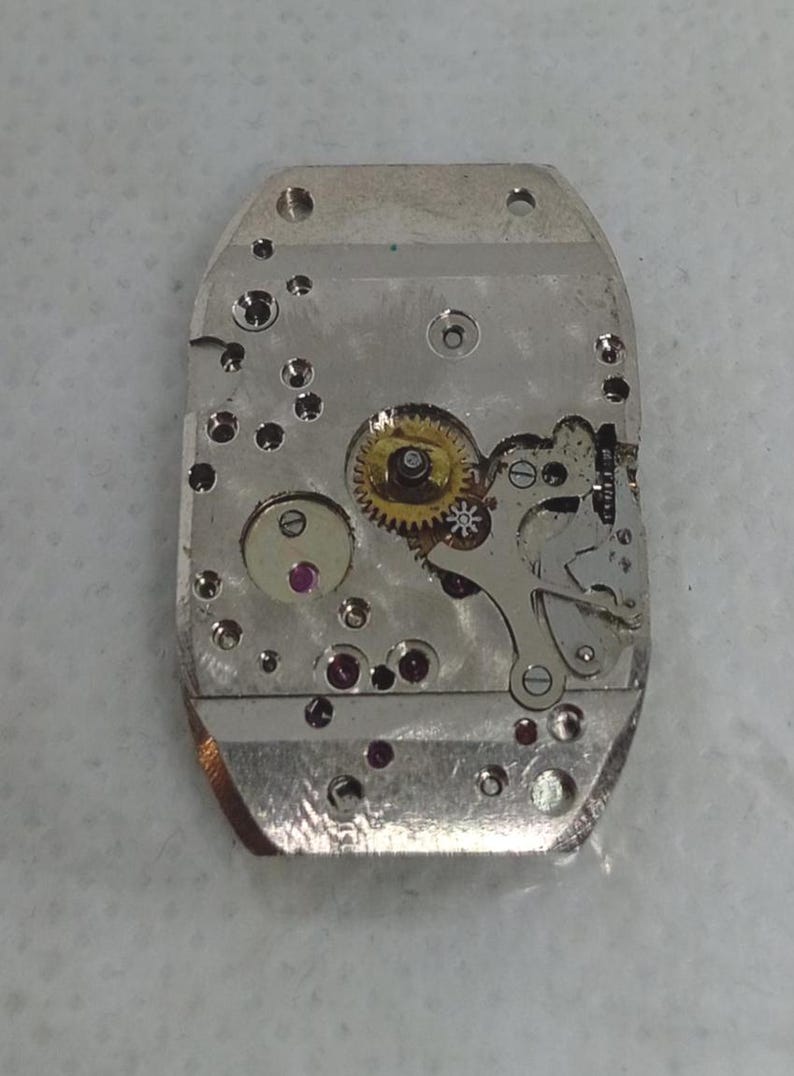 May include: Close-up of a watch movement, showcasing intricate gears, jewels, and metal components. The silver-colored base holds a complex arrangement of small parts, including a gold-colored gear and several red jewels. The overall design is rectangular with rounded edges.