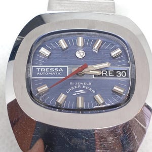 May include: A vintage Tressa Automatic watch with a blue face and silver-tone case. The watch features a rectangular face with silver markers and hands, and the text "RE 30" is visible. The watch band is silver-tone.