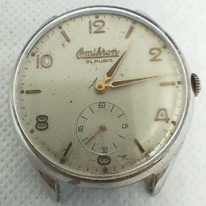 Könnte beinhalten: Eine Vintage-Omikron-Armbanduhr mit silberfarbenem Gehäuse und cremefarbenem Zifferblatt. Die Uhr hat goldfarbene Zeiger und Stundenmarkierungen sowie ein kleines Zifferblatt für die Sekunden. Der Text "Omikron" und "21 Rubis" ist auf dem Zifferblatt zu sehen.