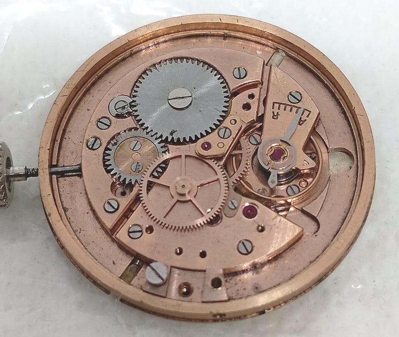 May include: Close-up of a watch movement with intricate gears, wheels, and ruby jewels. The metal components are in shades of gold and silver, showcasing the internal mechanics of a timepiece. The image highlights the precision engineering of the watch.