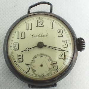 May include: An antique Cortébert wristwatch with a cream-colored face and black numerals. The watch has a small seconds dial and a dark metal case. The watch hands point to 4:10. The watch has a loop for a strap.