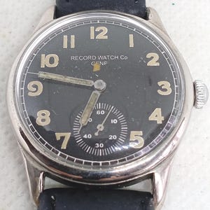 Op de afbeelding: Een zilverkleurig polshorloge met een zwarte wijzerplaat, voorzien van de tekst "RECORD WATCH Co GENE" in het wit. Het horloge heeft beige Arabische cijfers, een sub-wijzerplaat voor seconden onderaan en een zwarte leren band met een metalen kroon.