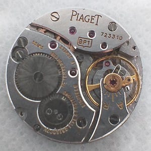 May include: Close-up of a silver Piaget watch movement with the brand name visible. The movement features intricate gears, jewels, and the text "SWISS". The model number 723310 is also visible.