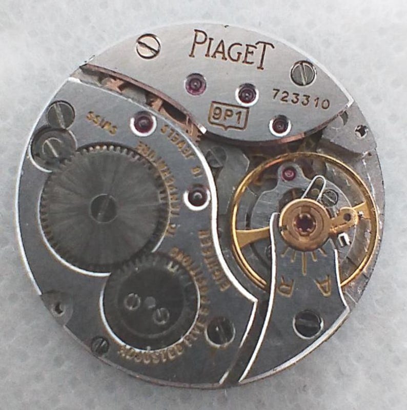 May include: Close-up of a silver Piaget watch movement with the brand name visible. The movement features intricate gears, jewels, and the text "SWISS". The model number 723310 is also visible.