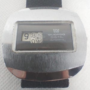 May include: A vintage Ducado Digital Automatic watch with a silver-toned case and a black band. The watch face displays the time with digital numerals. The watch has a rectangular display with the brand name and model details.